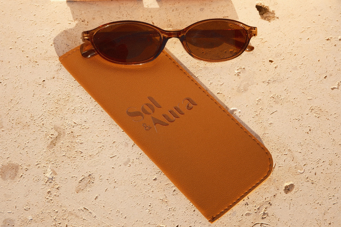 Brown sunglasses on a matching brown case with 'Sol & Aura' text, on a tiled surface.
