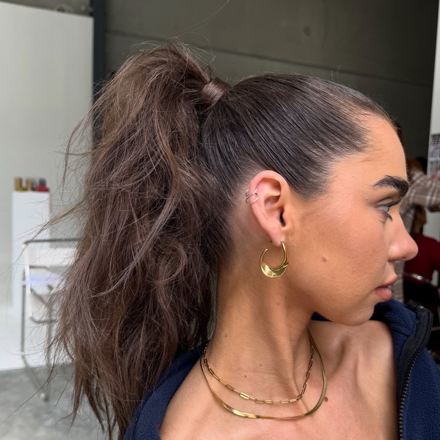 Woman with a high ponytail wearing gold hoop earrings and necklaces.