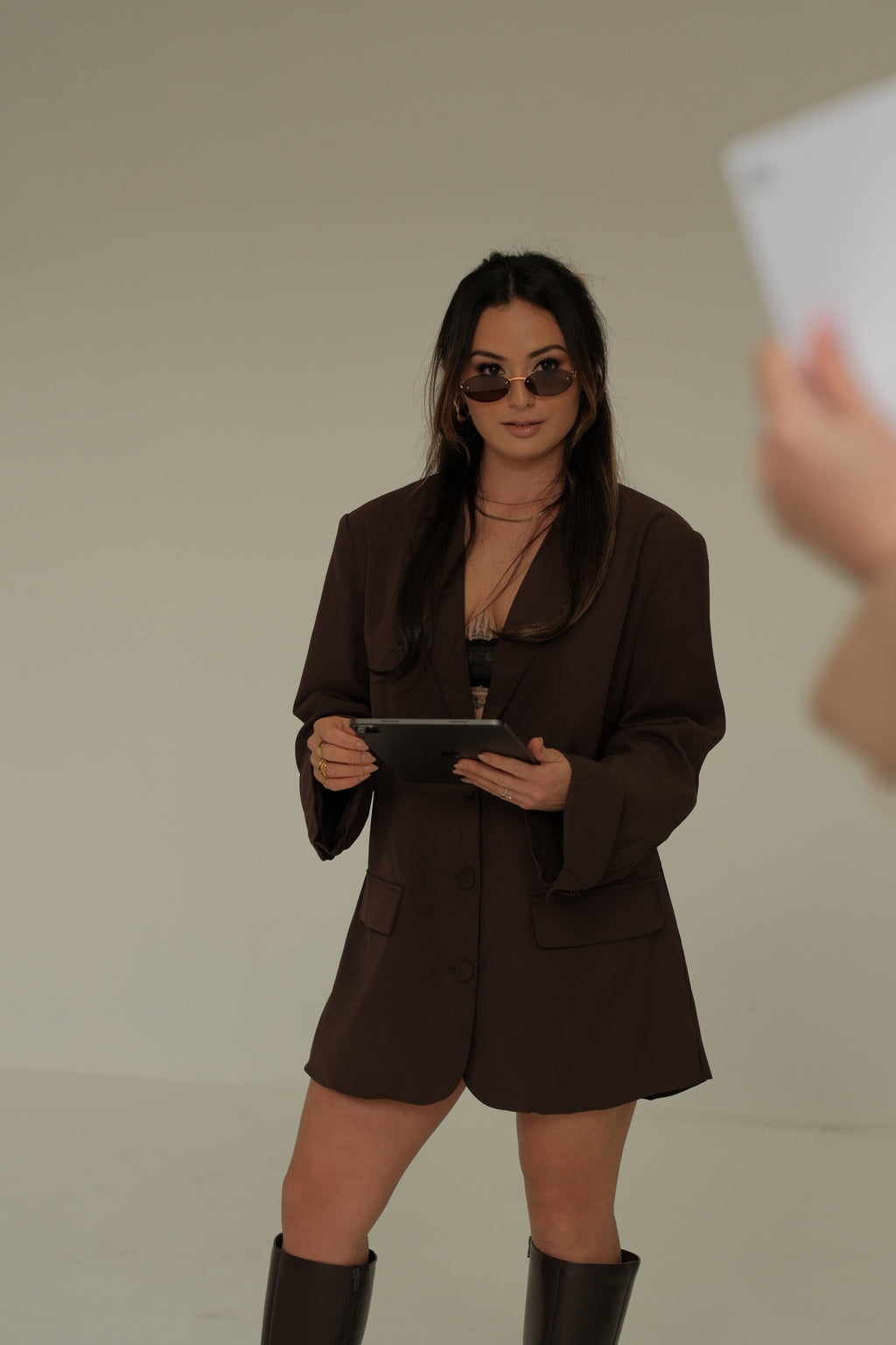 Woman in a brown outfit wearing sunglasses holding a tablet against a plain background