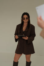 Woman in a brown outfit wearing sunglasses holding a tablet against a plain background