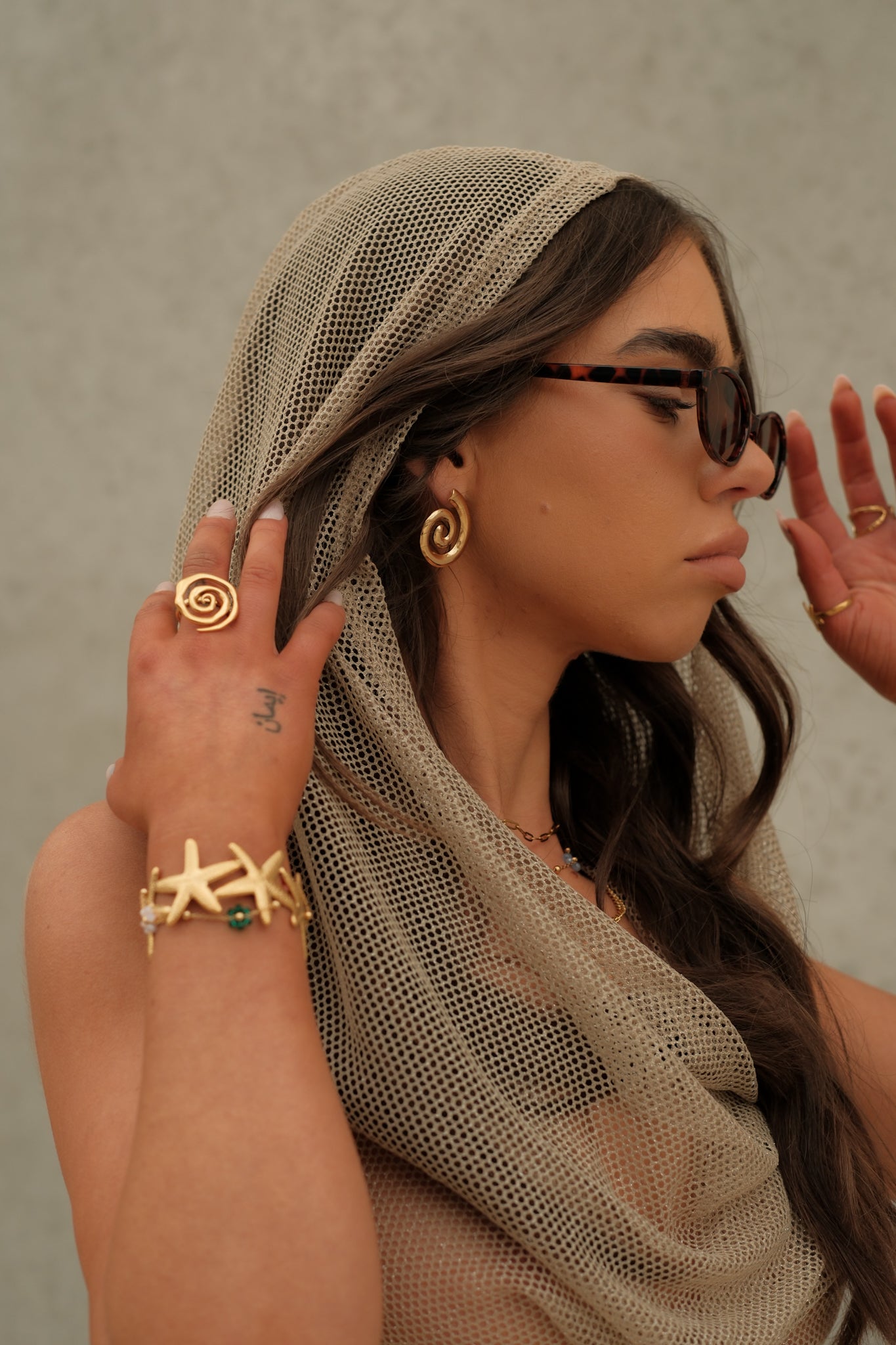 Woman wearing a beige headscarf, sunglasses, and gold jewelry against a neutral background