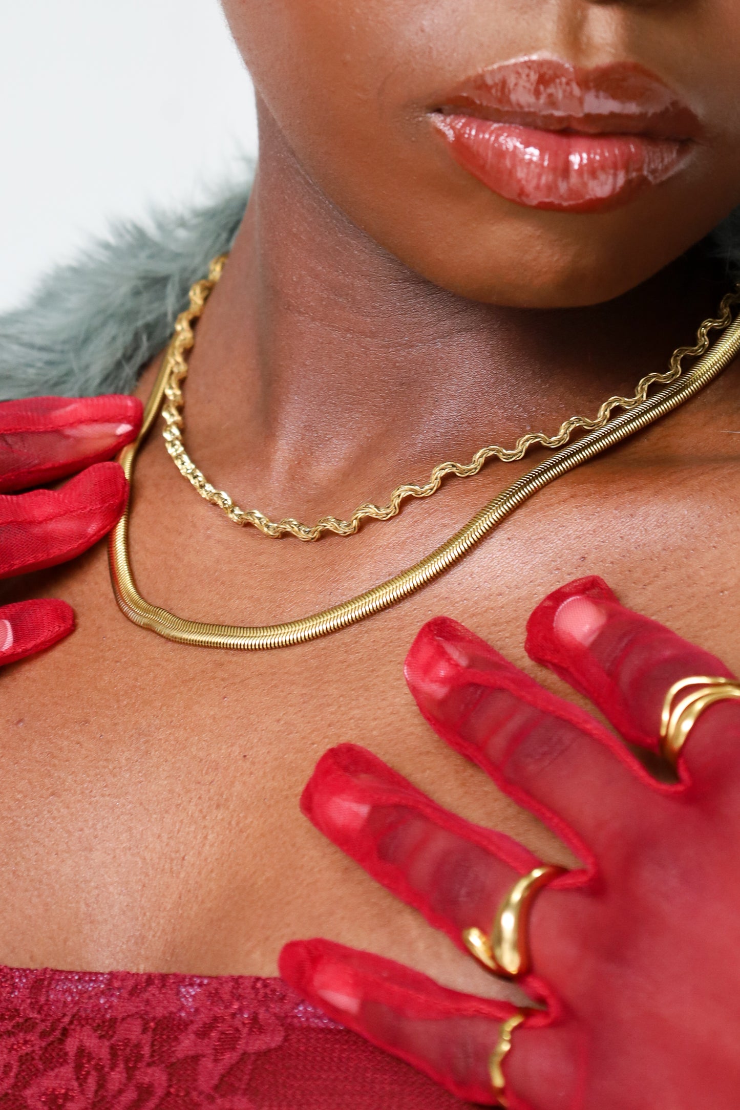 Close-up of a person wearing a gold plated waterproof hypoallergenic necklace and rings with red lace garment, red sheer gloves and green fur coat