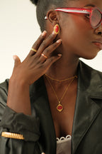 Woman wearing gold jewelry with red accents, including earrings and a necklace, against a neutral background.