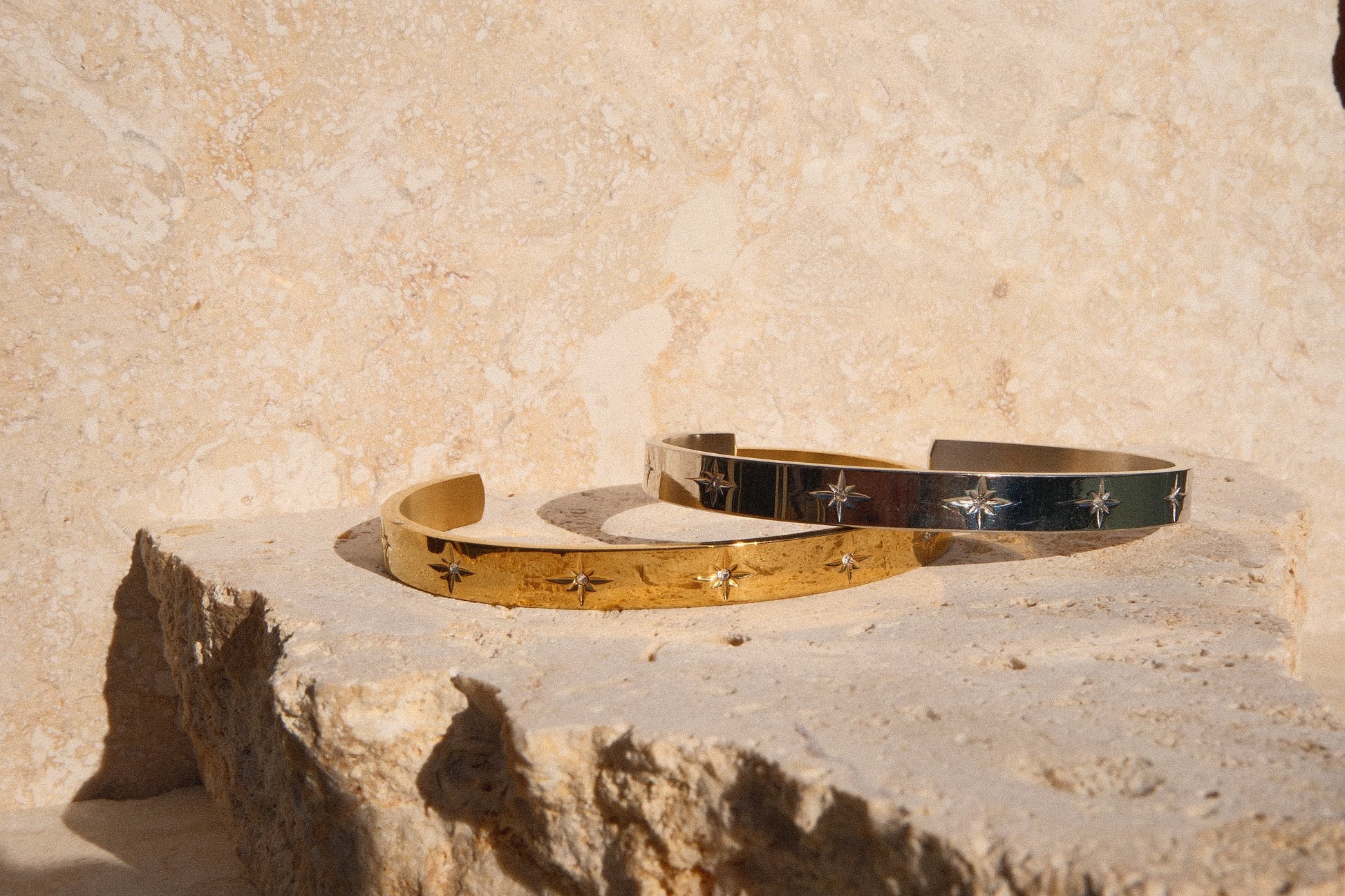 Two gold and one silver bracelet on a stone surface with a beige stone wall background