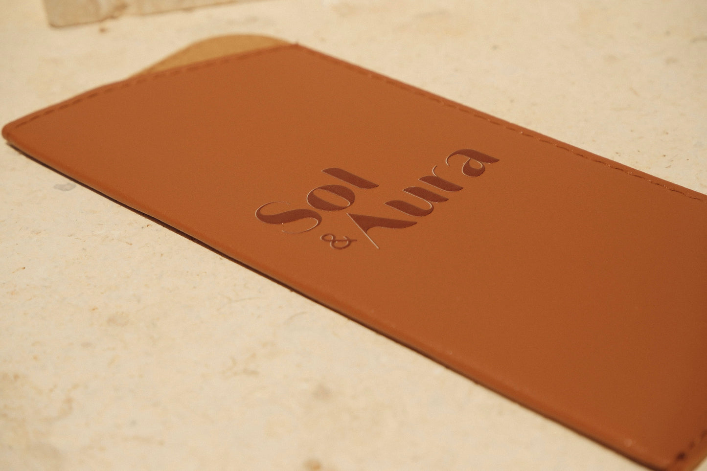 Brown leather sunglasses case with 'Sol & Aura' branding on a stone surface