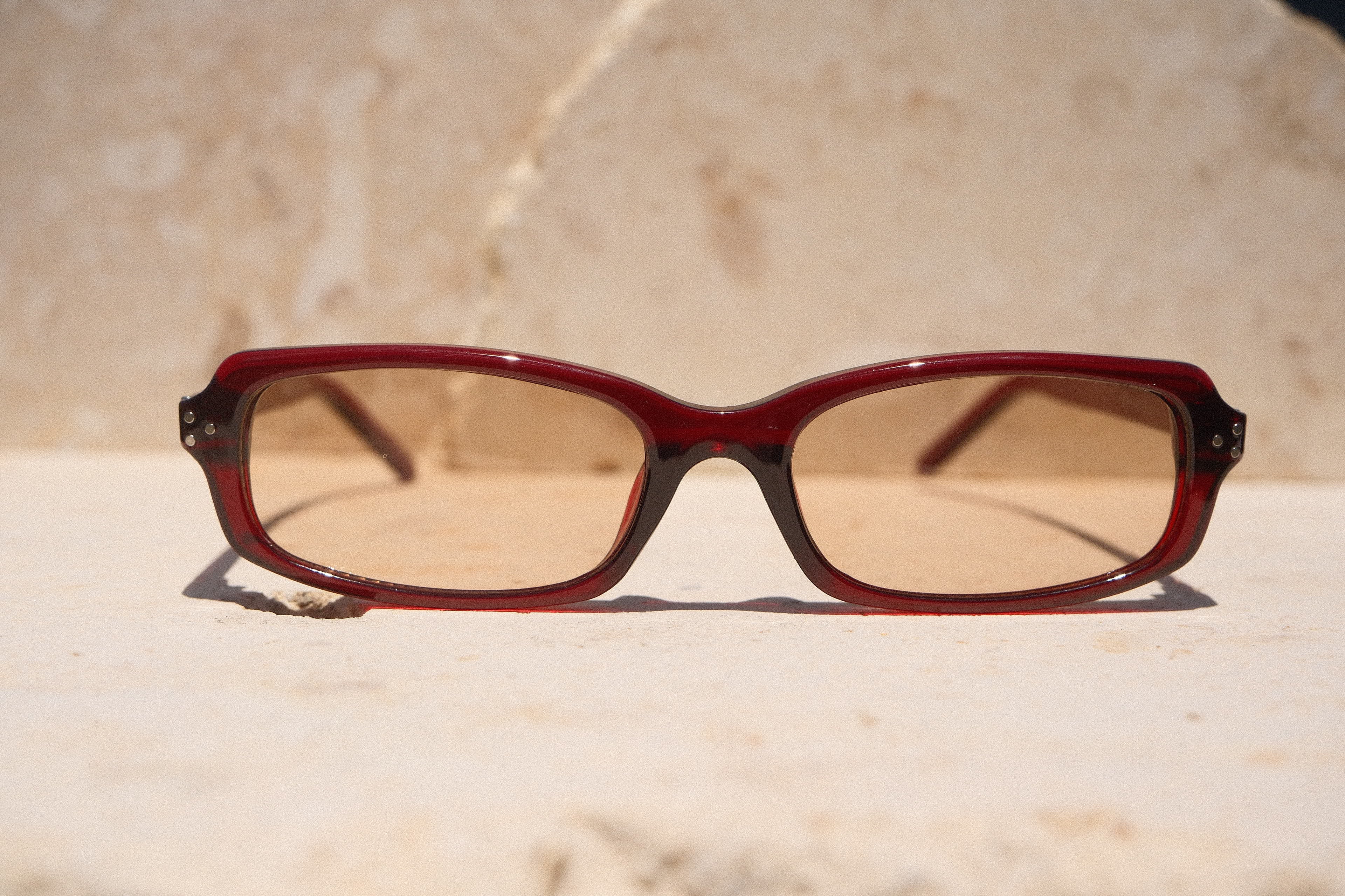 Sol & Aura - Erin Sunglasses. Red sunglasses with light tinted lens sitting on stone surface with three silver dot detailing on either side of lens