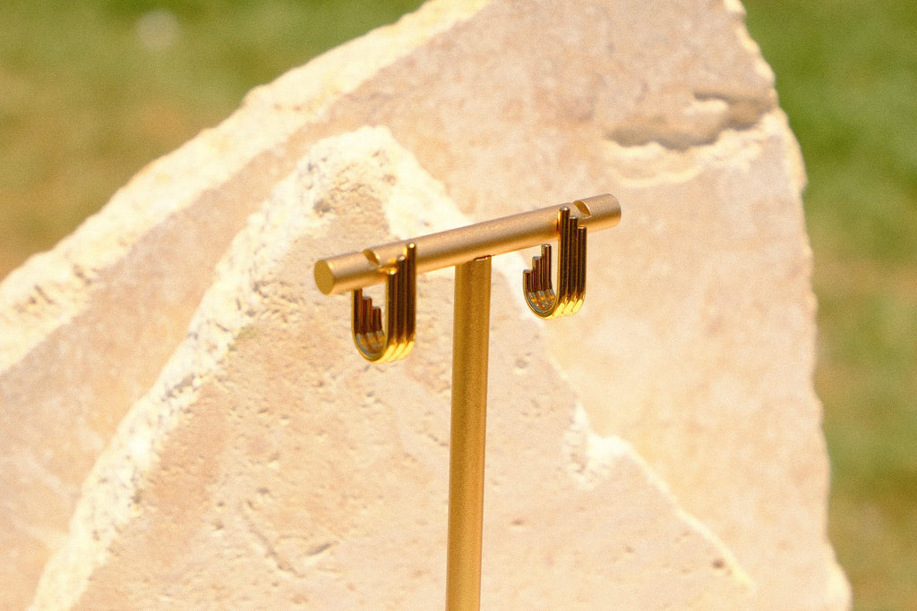 Sol & Aura - Gold stud earrings hanging on earring display against stone surface