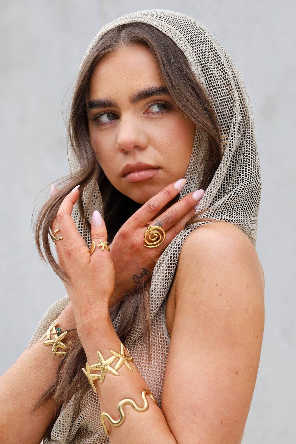 Woman wearing a mesh head covering and gold jewelry against a neutral background