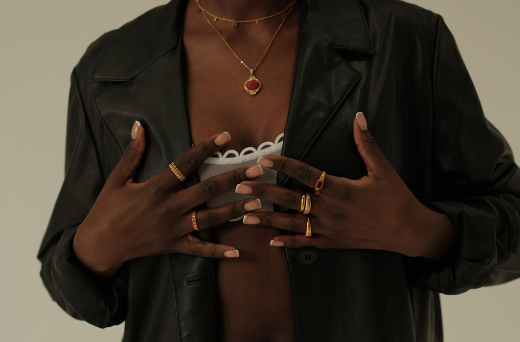 Person wearing a black jacket with gold jewelry, including rings and necklaces, on a neutral background.