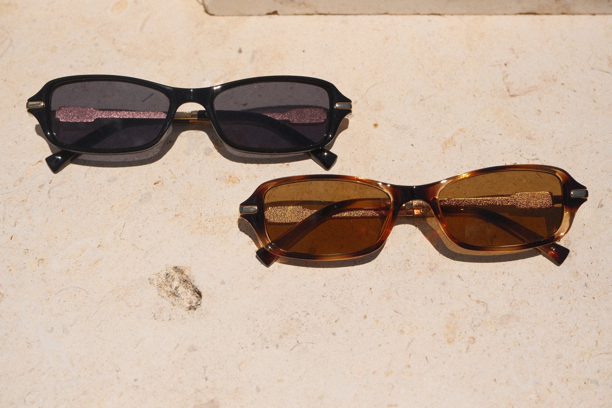 Sol & Aura - Jordan Sunglasses, black and brown with gold accents, lying on stone base