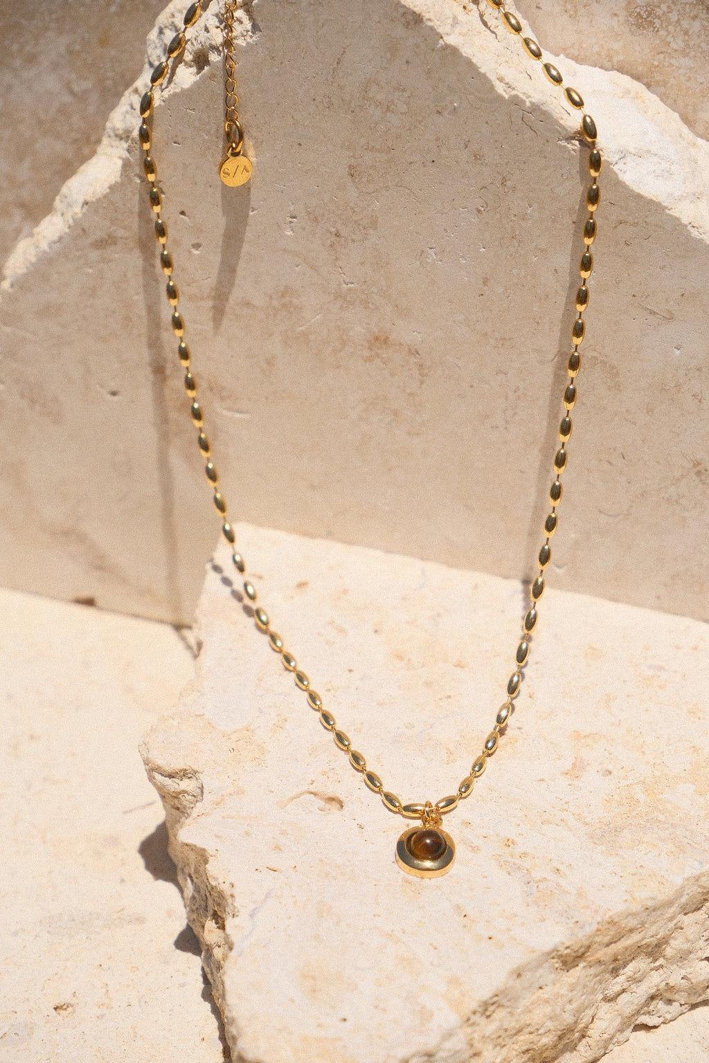 Sol & Aura - Minx Necklace. Brown pendant Tiger's Eye pendant on gold beaded chain lying on stone surface