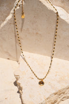 Sol & Aura - Minx Necklace. Brown pendant Tiger's Eye pendant on gold beaded chain lying on stone surface