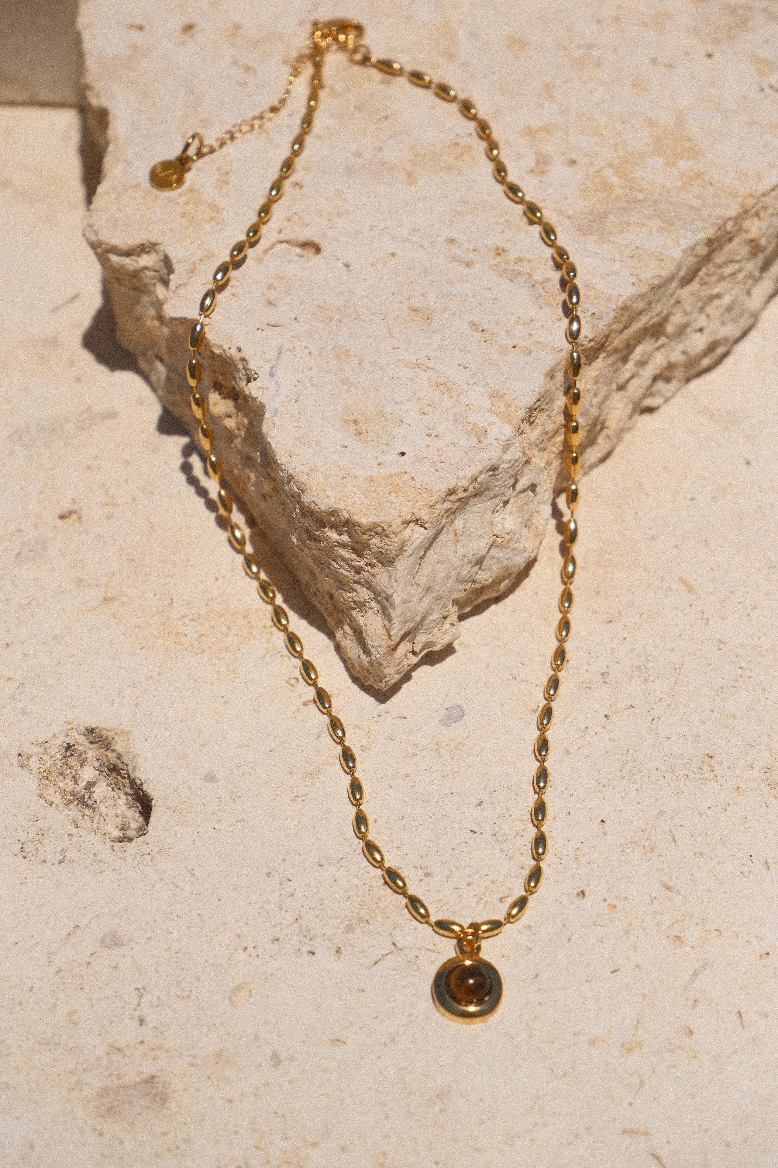 Sol & Aura - Minx Necklace. Brown pendant Tiger's Eye pendant on gold beaded chain lying on stone surface