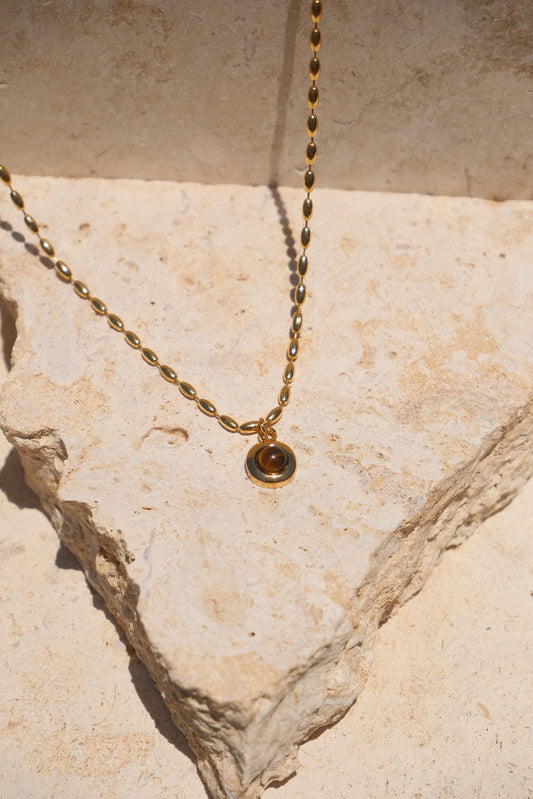 Sol & Aura - Minx Necklace. Brown pendant Tiger's Eye pendant on gold beaded chain lying on stone surface