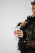 Person wearing a black lace garment and sunglasses, holding a glass against a white background
