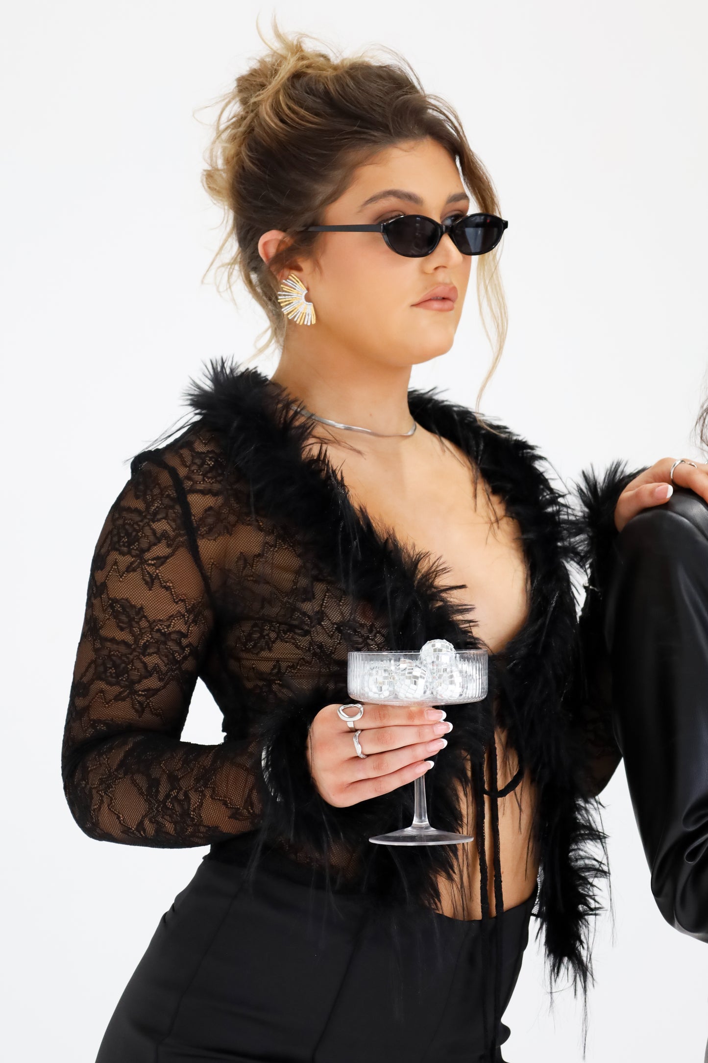 Sol & Aura - Woman wearing Sicily sunglasses in black, Oh Baby ring in silver and Ray earrings in black outfit with lace and fur trim. Woman is holding a glass filled with mini disco balls 