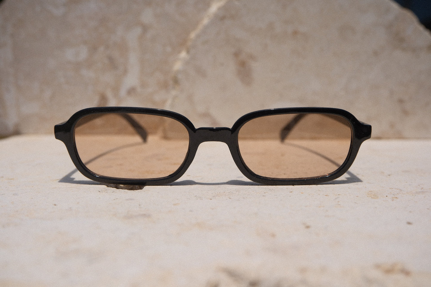 Sol & Aura - Carrie Sunglasses - black frame with tea-coloured lens. Narrow rectangular lens shape, front view on stone surface