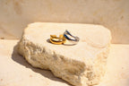 Sol & Aura - Two rings, one gold and one silver, on a textured stone surface with spiral sun designs