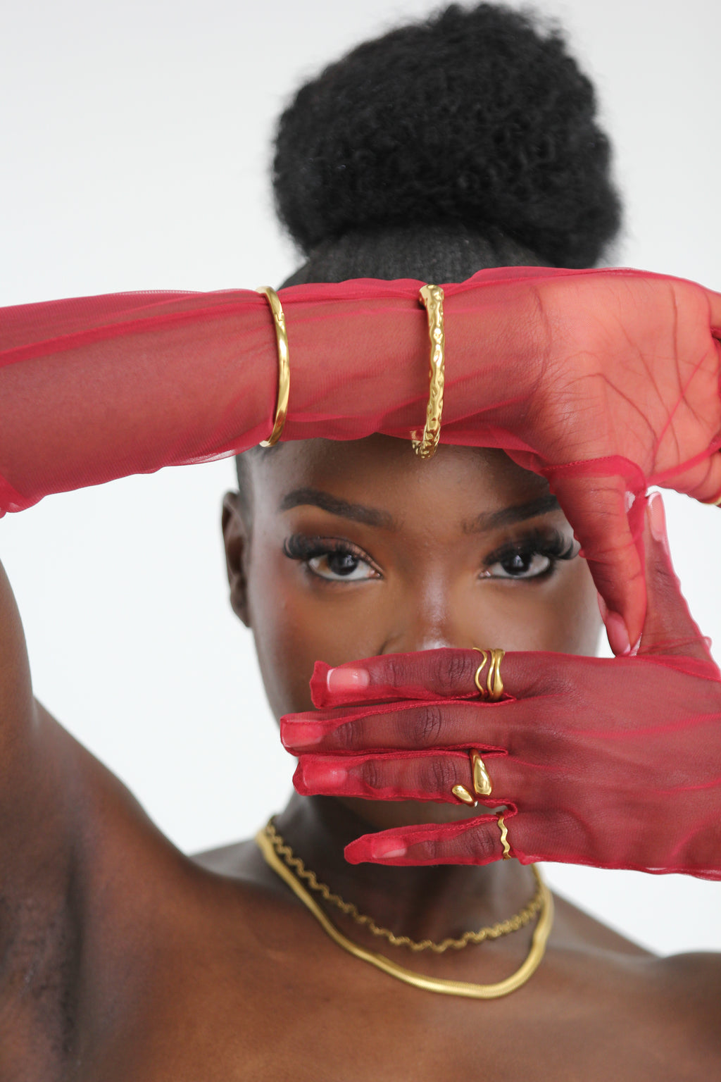 Sol & Aura - Muse Bangle, Ripple RIng, Reign Ring - Woman framing face with hands in red mesh gloves & gold Sol & Aura jewellery